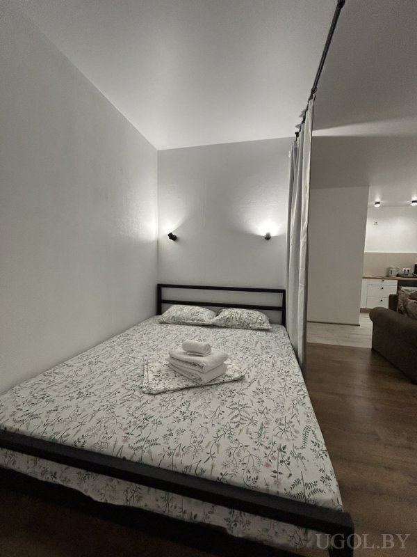 Кровать с белым покрывалом и зелёным ботаническим узором, настенные светильники, кухня на фоне и коричневый диван.