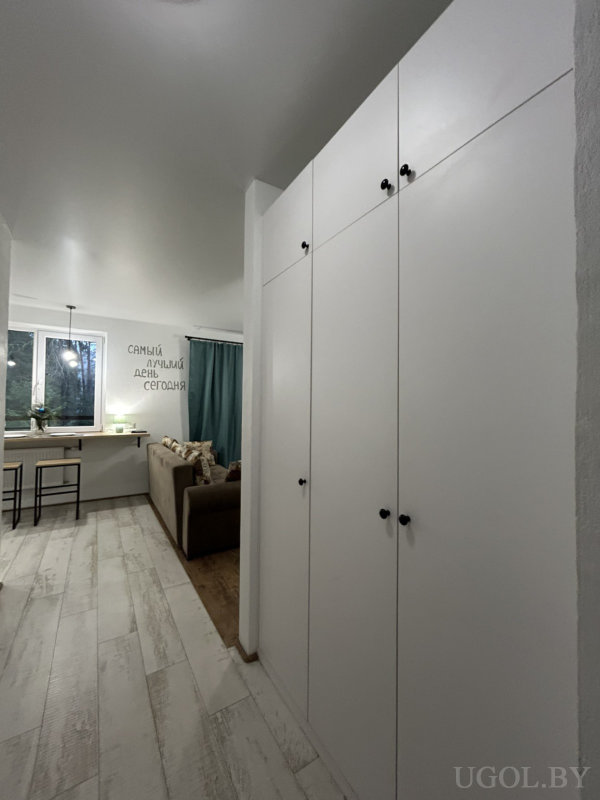 Светлая прихожая с высоким белым шкафом, деревянным полом и видом на гостиную с надписью «САМЫЙ ЛУЧШИЙ ДЕНЬ СЕГОДНЯ».