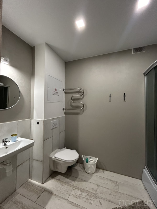 Современная ванная с подвесным унитазом, белой раковиной, круглым зеркалом и душевой кабиной с изогнутым стеклом.