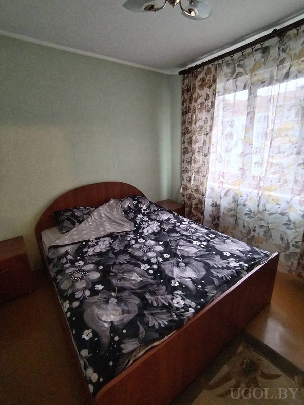 Небольшая спальня с деревянной кроватью, тёмным покрывалом с цветами, прикроватными тумбами и окном с цветочными шторами.