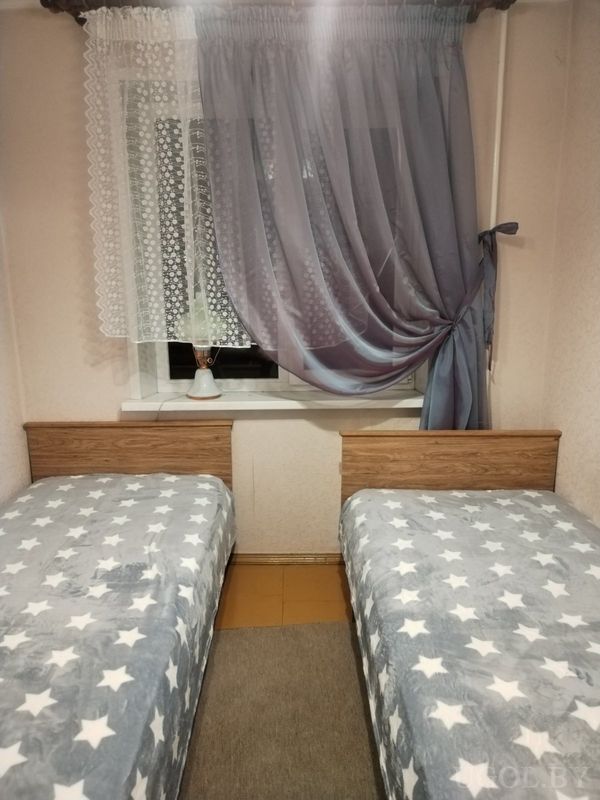 Компактная спальня с двумя односпальными кроватями, серыми покрывалами со звёздами, окном с белыми и серыми шторами и лампой на подоконнике.