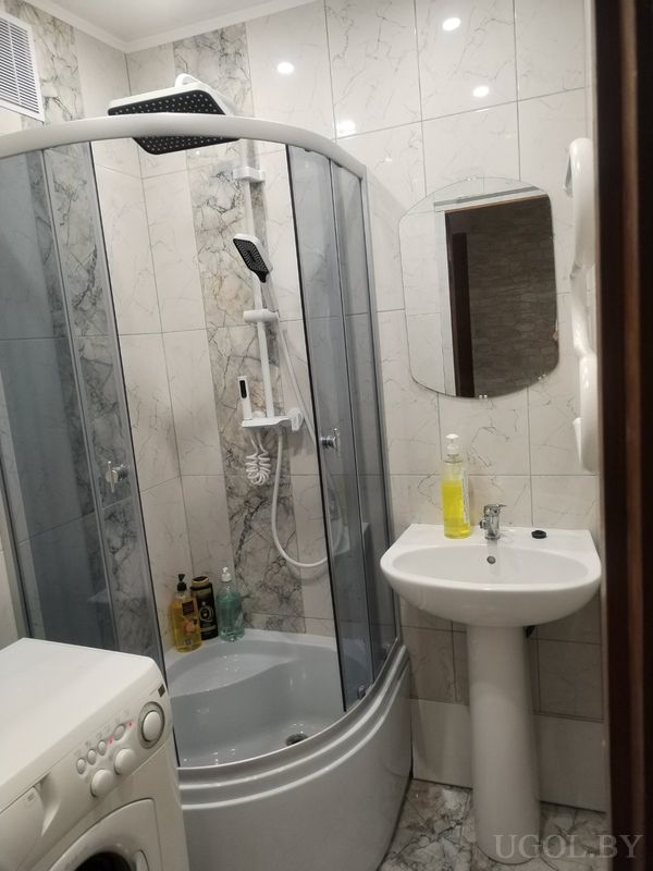 Компактная ванная с душевой кабиной, белой стиральной машиной, умывальником с зеркалом и мраморной плиткой.