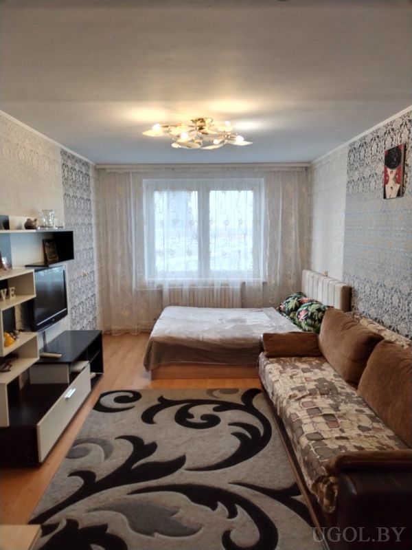 Комната с кроватью у окна, диваном с покрывалом, телевизором на светлой стенке и большим ковром с узором.