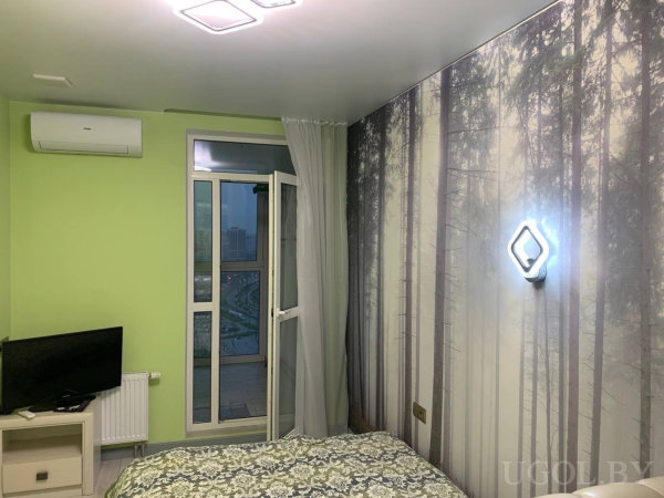 Комната с лесным панно, кондиционером, телевизором и выходом на балкон с городским пейзажем.