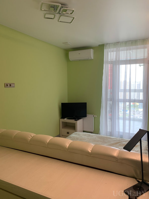 Светлая комната с белым диваном, телевизором, кондиционером и видом на город через большое окно.