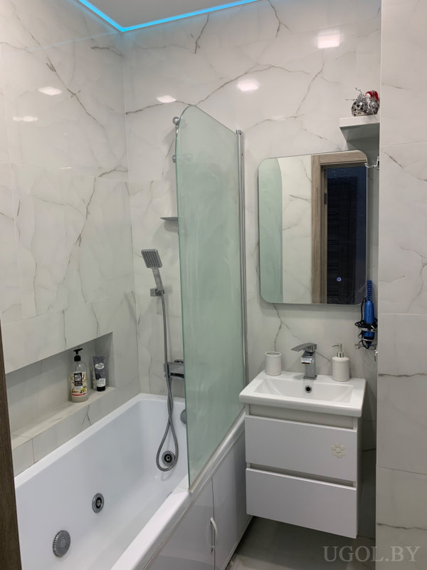 Интерьер с ванной, стеклянной перегородкой, белой тумбой и синим феном на стене.