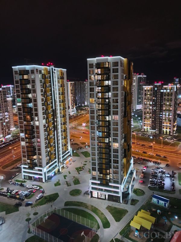 Панорама с высоты на две башни, освещённые фасады, спортивную площадку и дороги с машинами.