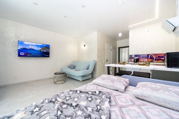Открытая студия с кроватью в звёздном текстиле, кухней с белой столешницей, голубым диваном и телевизором на стене.