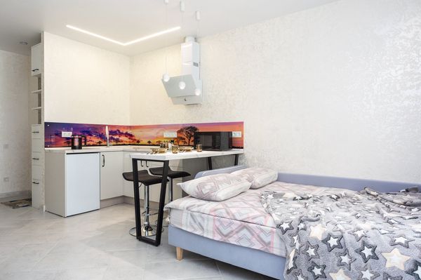 Компактная студия с кухней, фартуком с закатом, барной стойкой, двумя табуретами и кроватью с сиреневым каркасом.