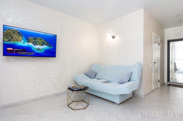 Минималистичная гостиная с голубым диваном, золотым столиком, телевизором и проходом в спальню.