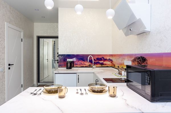 Кухня с белыми шкафами, фартуком с закатом, вытяжкой, кофемашиной и сервировкой на мраморной столешнице.