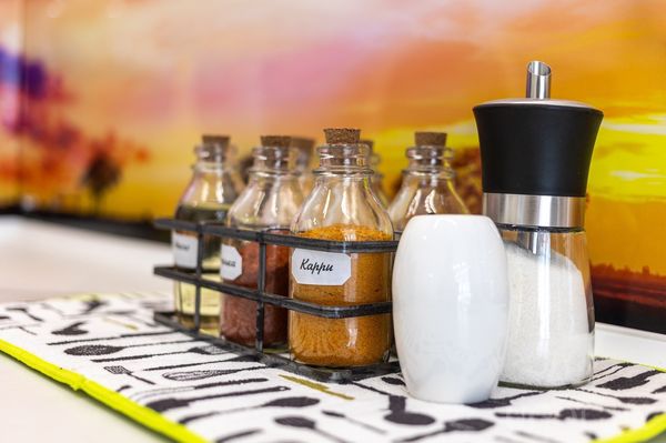 Кухонная полка с банками специй, контейнерами и фартуком с тёплым пейзажем на фоне.