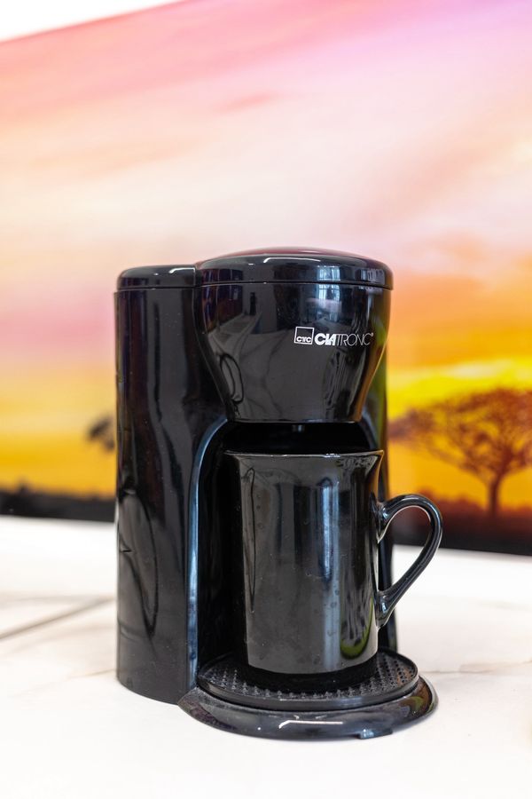 Чёрная кофемашина Clatronic с кружкой на белой столешнице, на фоне фартука с закатным пейзажем.