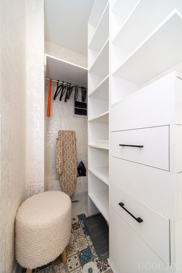 Хозяйственная комната с белыми полками, гладильной доской, вешалками и цветным плиточным полом.