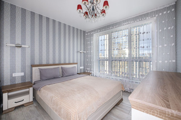 Кровать с бежевым покрывалом и серыми подушками, две тумбы, полосатые обои, большое окно с тюлем и красной люстрой, вид на городские высотки.