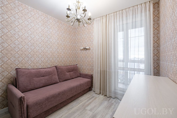 Сиреневый диван с подушками у стены с геометричными обоями, рядом стол у большого окна с тюлем и люстра с несколькими лампами.