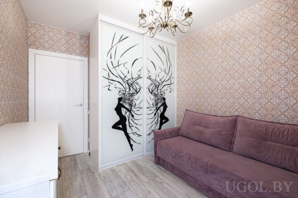 Сиреневый диван, белый стол и шкаф‑купе с декоративными силуэтами на дверцах, светлые обои и люстра создают уютный современный интерьер.