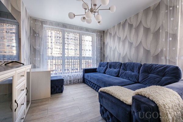 Синий диван с пледом, белая тумба с телевизором, рабочий стол с пуфом у окна с тюлем и вид на городские высотки.