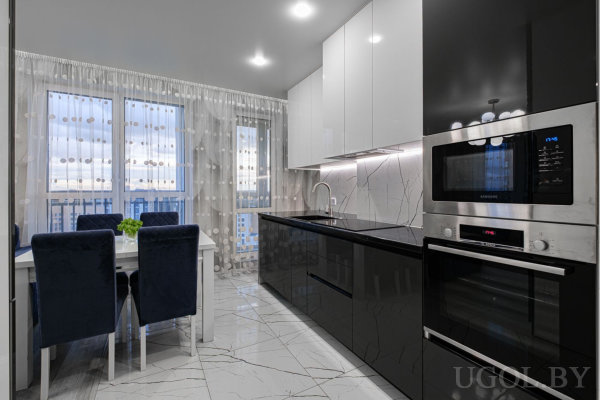 Кухня с белыми верхними и чёрными нижними фасадами, мраморным фартуком, встроенной техникой и обеденным столом с синими стульями у окна.