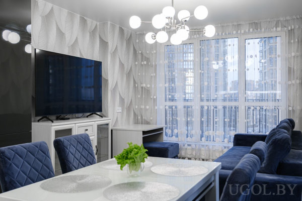 Обеденный стол с синими стульями, телевизор на белой тумбе у стены с волнистым рисунком, рядом синий диван и большие окна с тюлем.