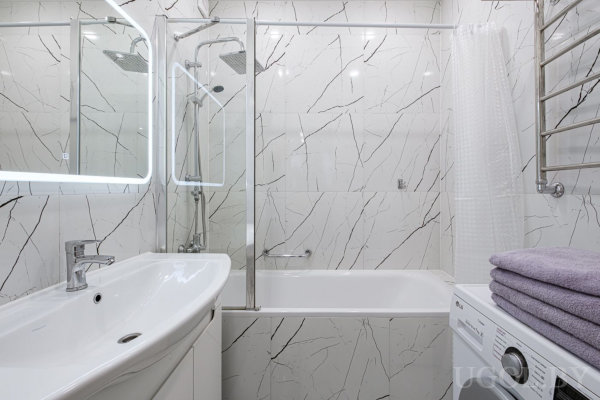 Современная ванная с мраморными стенами, ванной со стеклянной перегородкой, раковиной с подсвеченным зеркалом и стиральной машиной.