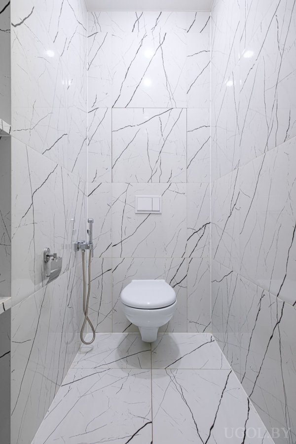 Узкий санузел с подвесным унитазом, мраморной плиткой, гигиеническим душем и скрытой системой смыва.