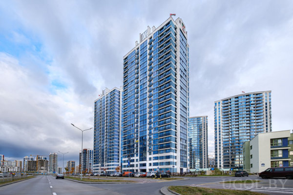 Городской вид на группу современных жилых башен с синими стеклянными фасадами, парковкой и широкой дорогой на переднем плане.