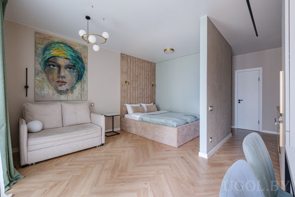Светлая кровать у акцентной стены, рядом бежевый диван и яркая картина, деревянный пол ёлочкой и современная люстра.