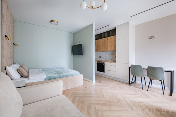 Кровать с бело-зелёным бельём, рядом диван, кухня с деревянными и белыми шкафами, обеденный стол и настенный ТВ.