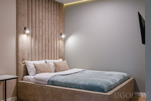 Кровать с голубым покрывалом и светлыми подушками, мягкое освещение, серый фон с панелями и настенный телевизор.