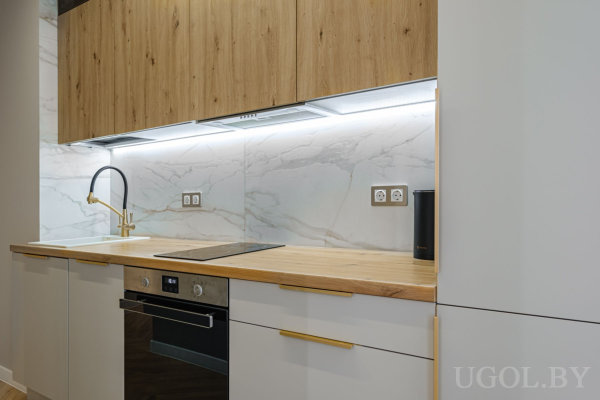 Белые нижние шкафы с золотыми ручками, деревянная столешница, встроенная плита и духовой шкаф, мраморный фартук и золотой смеситель.