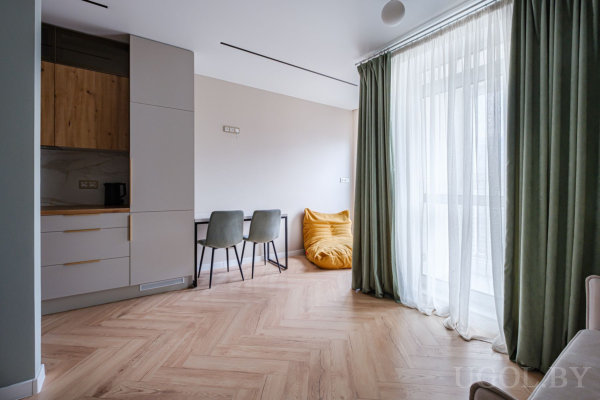 Компактная кухня с белыми и деревянными шкафами, стол с зелёными креслами, жёлтый пуф у окна и большие панорамные шторы.