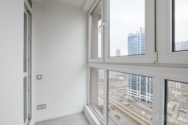 Застеклённая лоджия с полом и стенами светлого тона, вид на многоэтажные дома и дорогу с машинами.