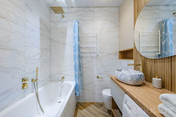 Белая ванна с золотым душем, туалет, деревянная тумба с мраморной раковиной, круглое зеркало и стиральная машина.