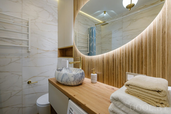 Круглое зеркало с подсветкой, деревянная столешница с мраморной раковиной и золотым смесителем, рядом туалет и полотенцесушитель.