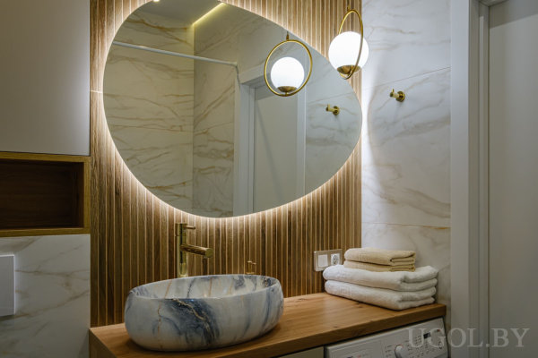 Зеркало с подсветкой на деревянной стене, мраморная раковина с золотым смесителем, деревянная столешница и стиральная машина.