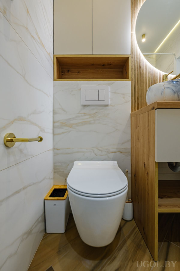 Настенный унитаз с скрытым бачком, золотой держатель бумаги, деревянная полка и шкаф, рядом часть тумбы с круглым зеркалом.