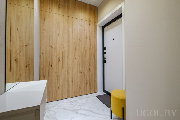 Белая дверь с чёрной рамкой, деревянные панели на стене, белые плитки на полу и жёлтый пуф рядом.