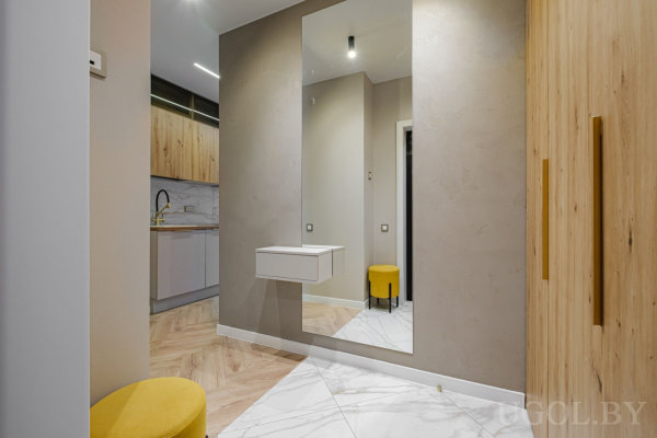 Большое зеркало с подвесной полкой, жёлтый пуф, переход к кухне с деревянными шкафами и мраморным фартуком.