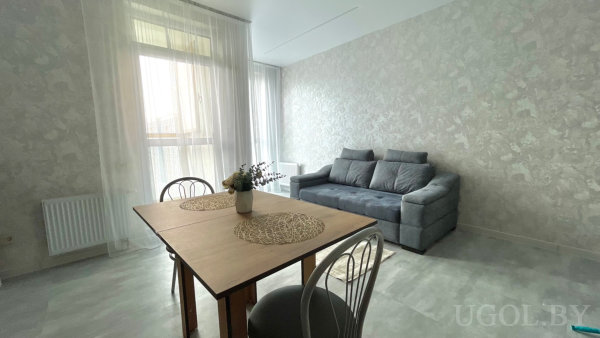 Деревянный стол с плетёными салфетками и вазой, металлические стулья и серый диван на фоне лёгких штор.