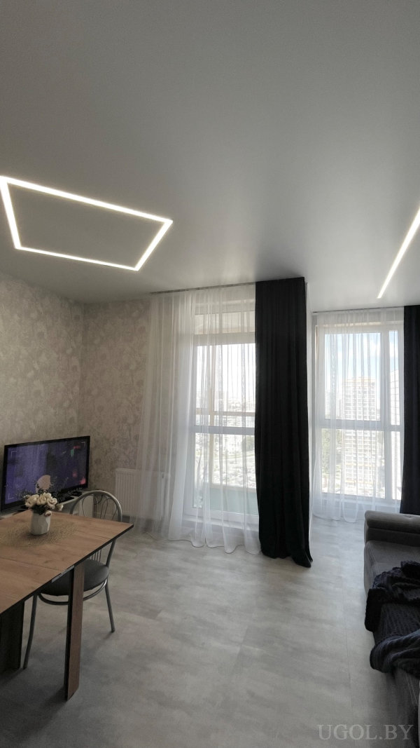 Напольные окна с тюлем и плотными шторами, прямоугольные LED-светильники, стол у окна и телевизор на стене.