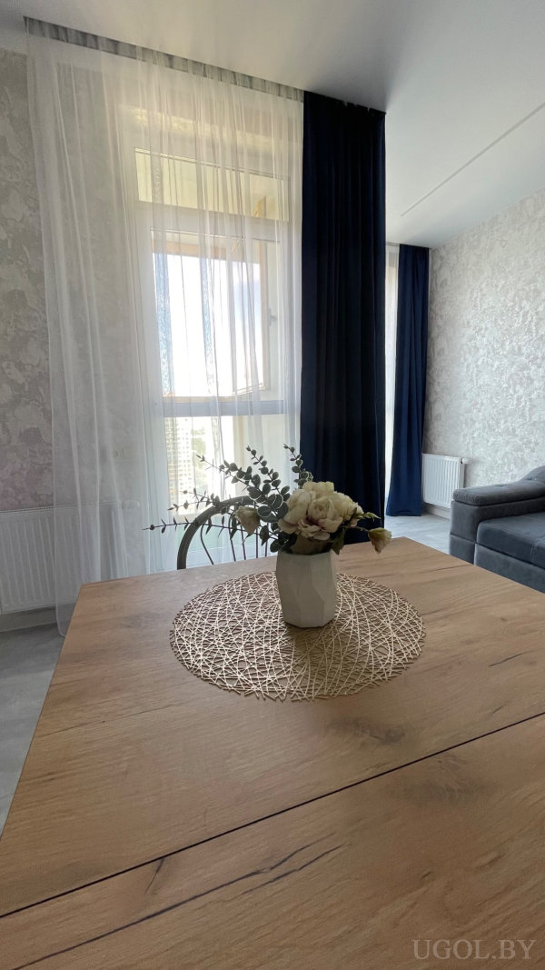 Белая ваза с бело-розовыми цветами на плетёной салфетке, серый диван и светлые стены с текстурой.