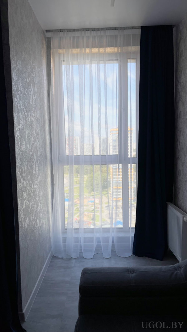 Полноразмерное окно с тюлем и плотными шторами, городской пейзаж с высотками и зелёной зоной внизу.