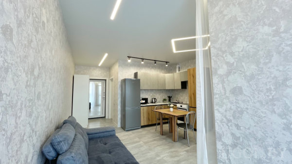 Открытая планировка с светло-деревянными и серыми фасадами, холодильник, микроволновка, стол на двоих и тёмно-серый диван.