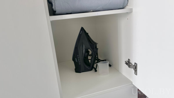 Белый шкаф с полками: снизу утюг и мерный стакан, сверху сложенное одеяло с голубым чехлом.