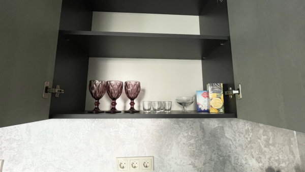 Открытые полки с бокалами, стаканами и десертной чашей, рядом коробка с лимонным соком и блок розеток на стене.