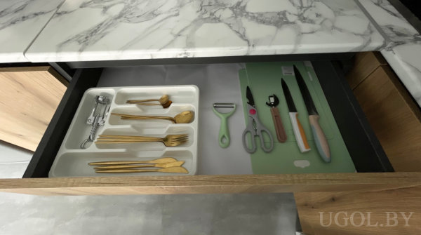 Лоток с золотистыми столовыми приборами и набором ножей, рядом овощечистка и ножницы на зелёном коврике.