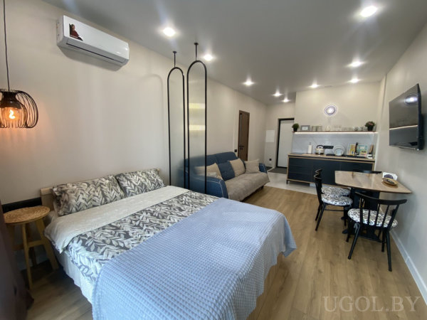 Кровать с голубым пледом, диван за декоративной перегородкой, кухня с открытыми полками и стол на четыре места.