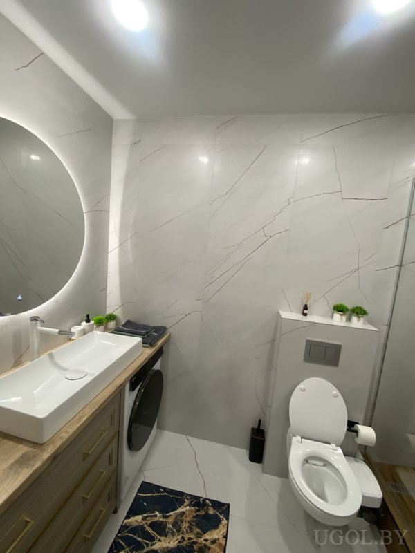 Раковина с круглым зеркалом, стиральная машина, туалет с декором, белая плитка и синий коврик.