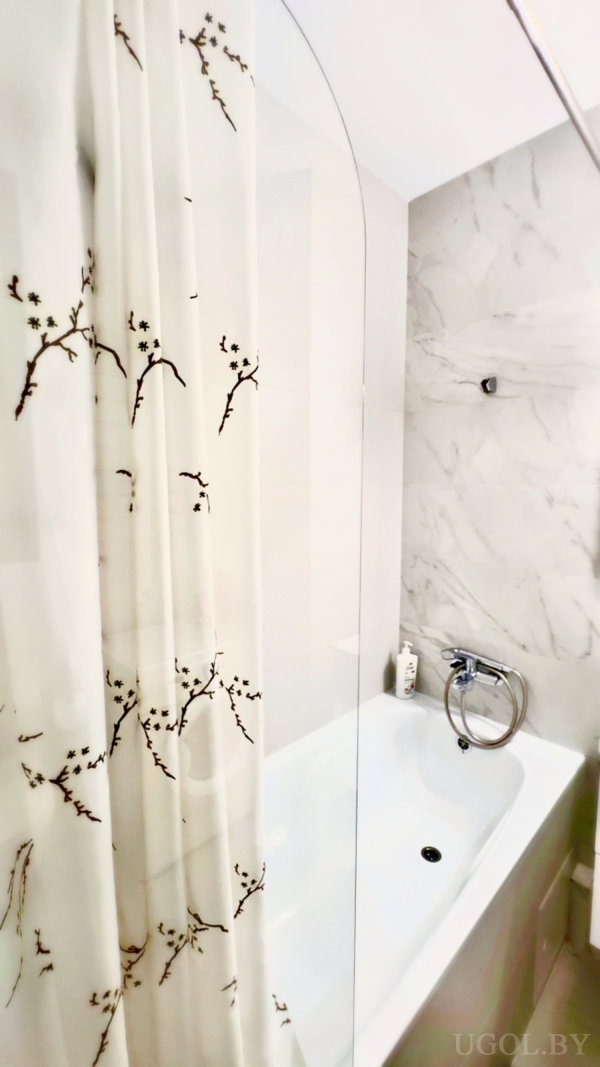 Комната с ванной, стеклянным экраном, душем, флаконом и белой плиткой с прожилками.